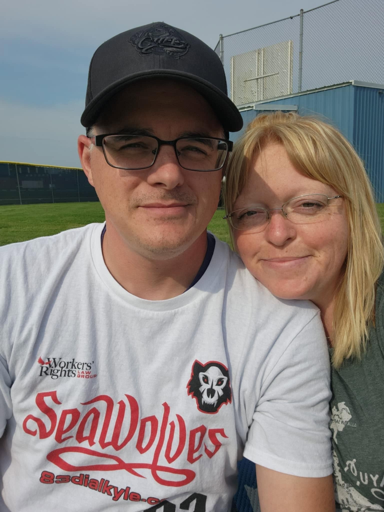 Smiling man in a SeaWolves shirt and blonde woman posing together at a baseball field.